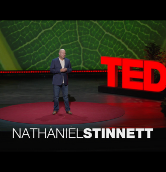 Nathaniel Stinnett stands on a stage in front of a large red sign that reads TED