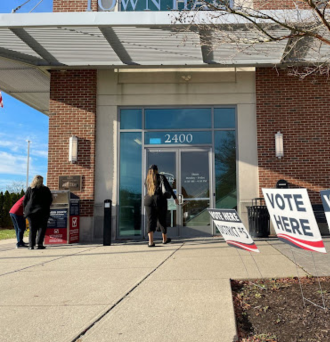 Three people outside of a polling place. Two people are using a drop box and one is walking into the building.